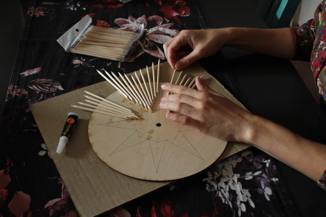 Ręce układają patyczki na okrągłej desce, tworząc wzór. To początek pracy nad lustrem okrągłym na pasku skórzanym jak zrobić samemu.