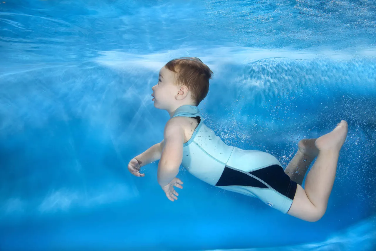Małe dziecko w jasnoniebieskim kombinezonie do pływania dla dzieci nurkuje w basenie, otoczone bąbelkami i światłem.