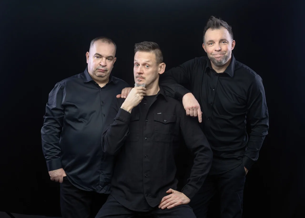 Trzech członk&oacute;w kabaretu Ani Mru Mru w czarnych koszulach pozuje do zdjęcia na czarnym tle.