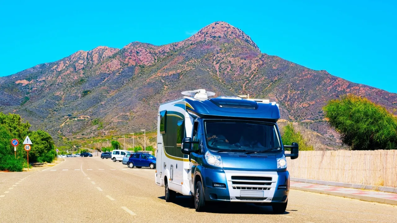 Niebiesko-biały kamper jedzie drogą w kierunku g&oacute;r. Idealny pojazd na podr&oacute;ż kamperem do Włoch.