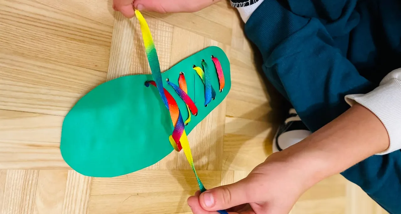 Dziecko ćwiczy, jak nauczyć dziecko wiązać buty, przewlekając tęczową sznurówkę przez zielony szablon buta.