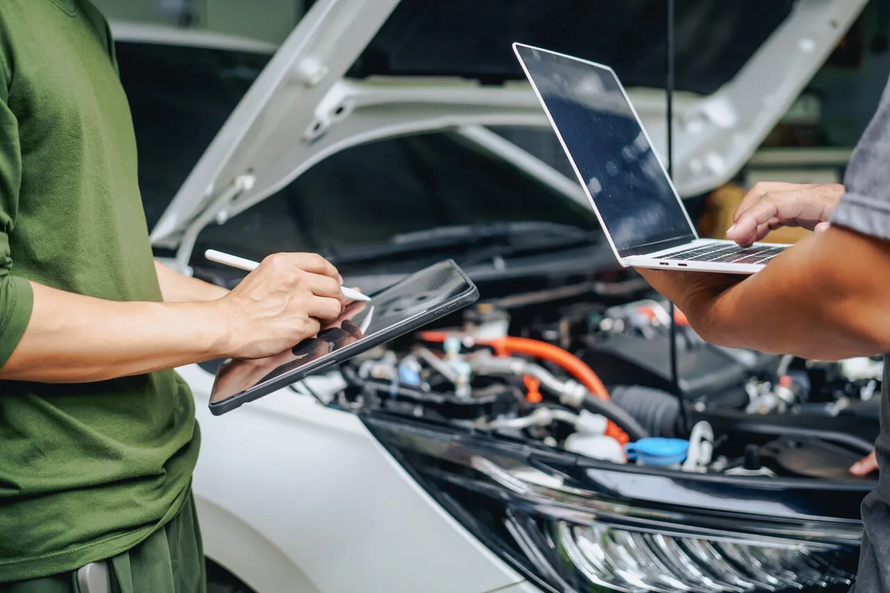 Mechanicy sprawdzają dane na tablecie i laptopie przy otwartej masce samochodu elektrycznego. Zastanawiają się, ile to km.