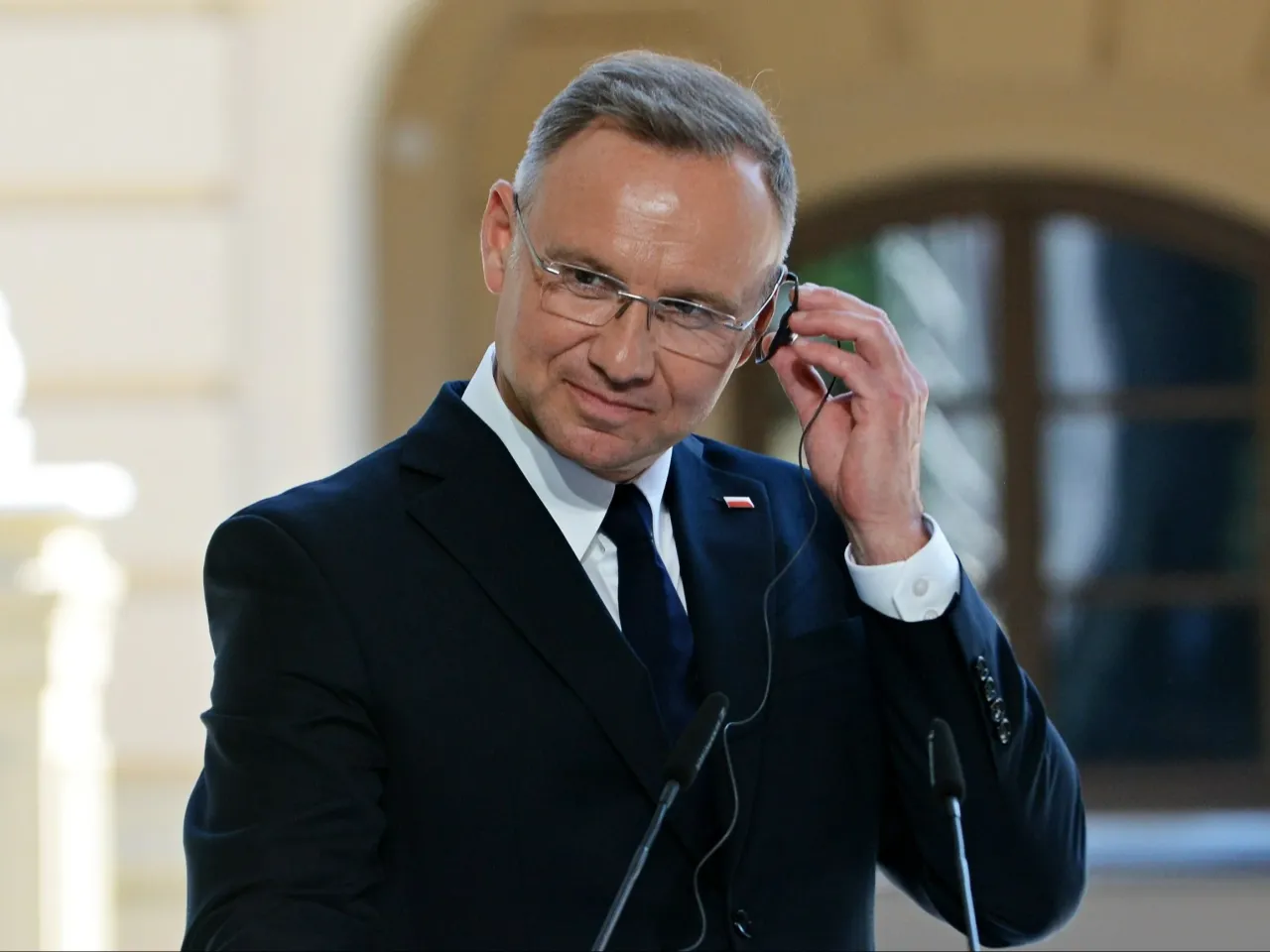 Prezydent Andrzej Duda w garniturze, z mikrofonem przy uchu. Jego wzrost jest imponujący, co widać po sylwetce.