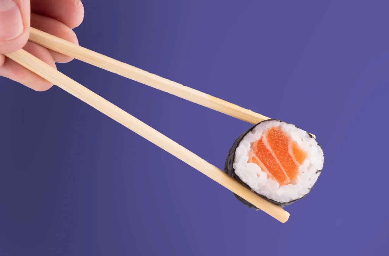 Dłoń pokazuje, jak trzymać pałeczki do sushi, by złapać kawałek rolki z łososiem.