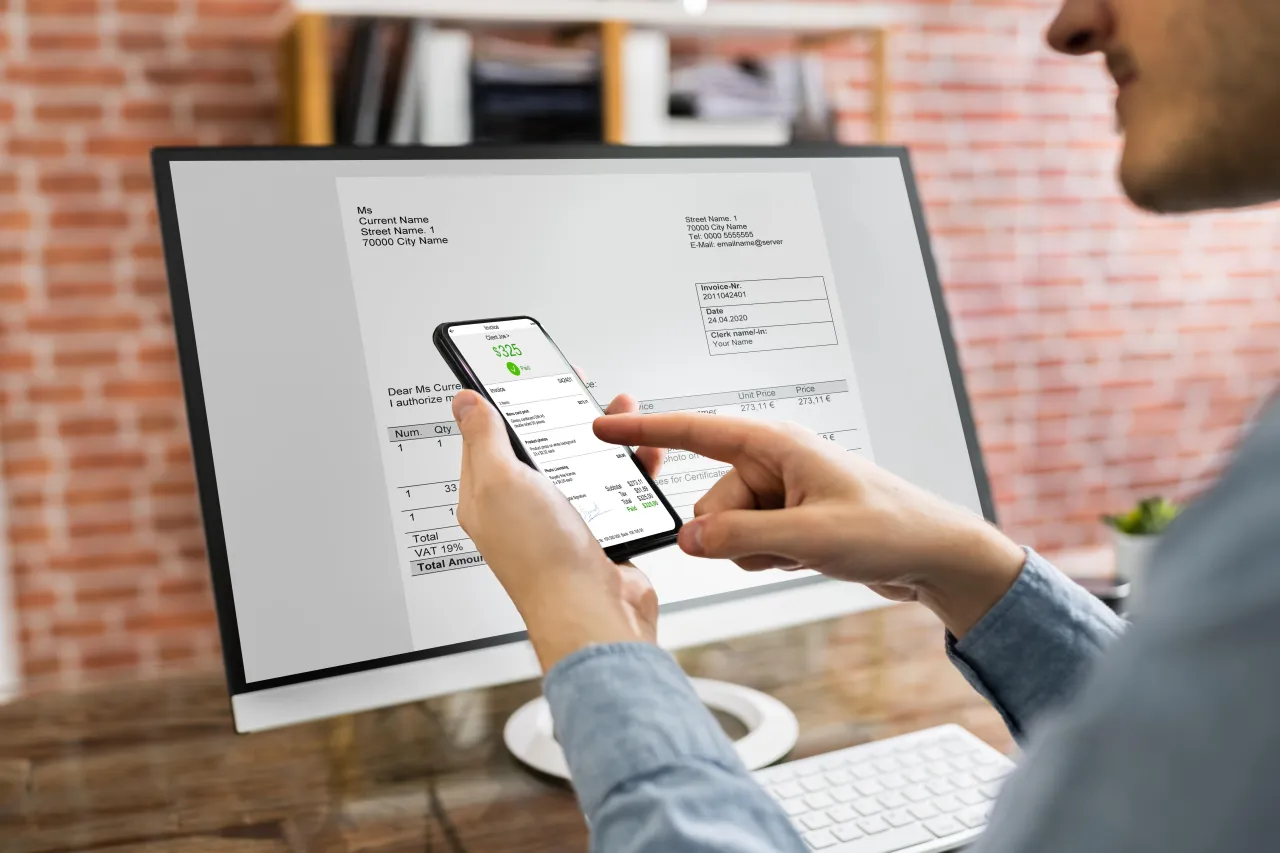 Osoba sprawdza na telefonie, jaki termin płatności na fakturze widocznej na monitorze.
