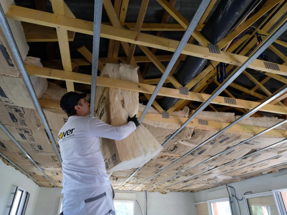 Pracownik montuje wełnę mineralną na suficie, izolując przewody elektryczne.