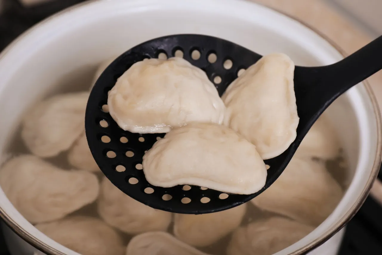 Gotowanie mrożonych pierog&oacute;w: na łyżce cedzakowej trzy gotowe pierogi, w tle garnek z gotującymi się pierogami.