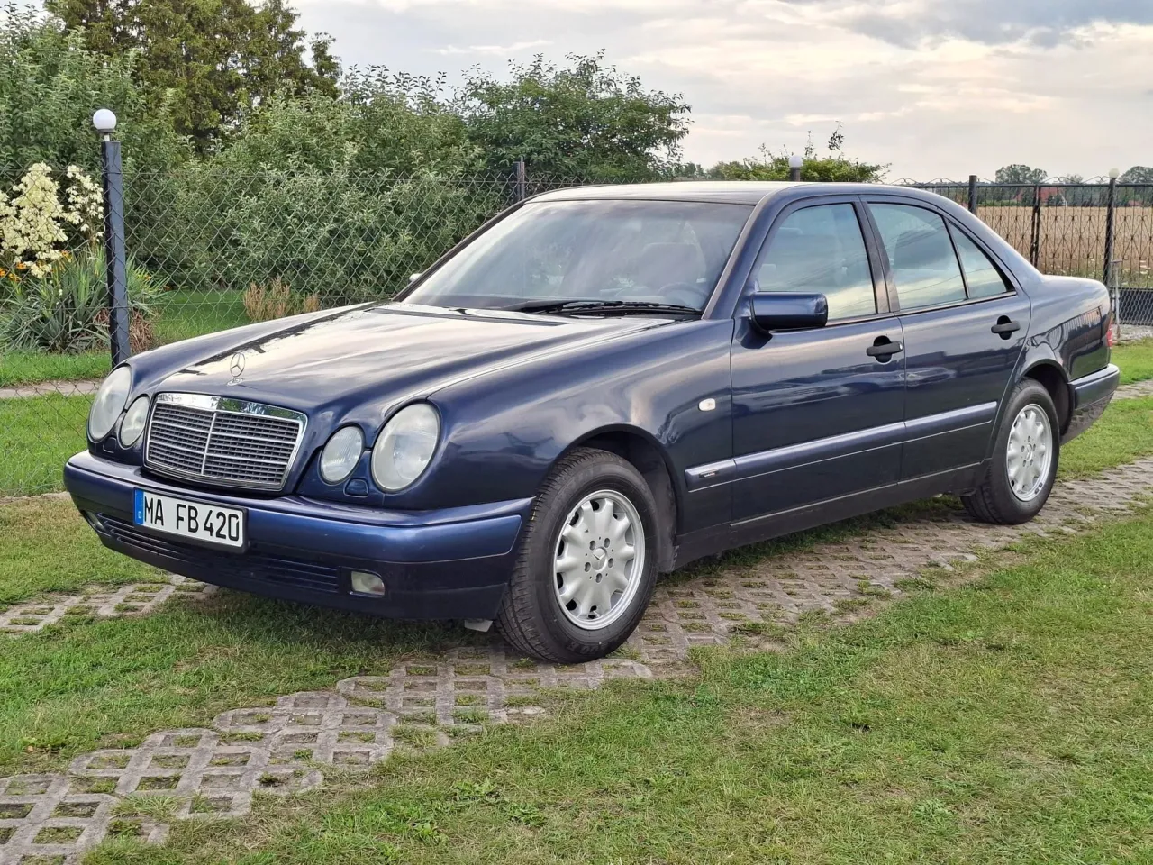 Granatowy Mercedes E klasa W210 z lat 90. stoi na trawniku obok betonowych płyt.