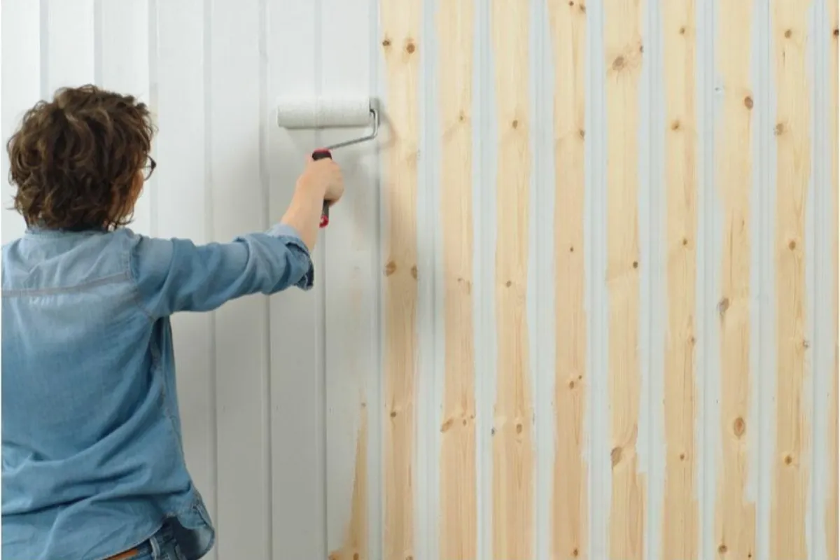 Osoba w dżinsowej koszuli maluje boazerię wałkiem. Widać, jak zmienia się kolor drewna, co pokazuje, jak pomalować boazerię.