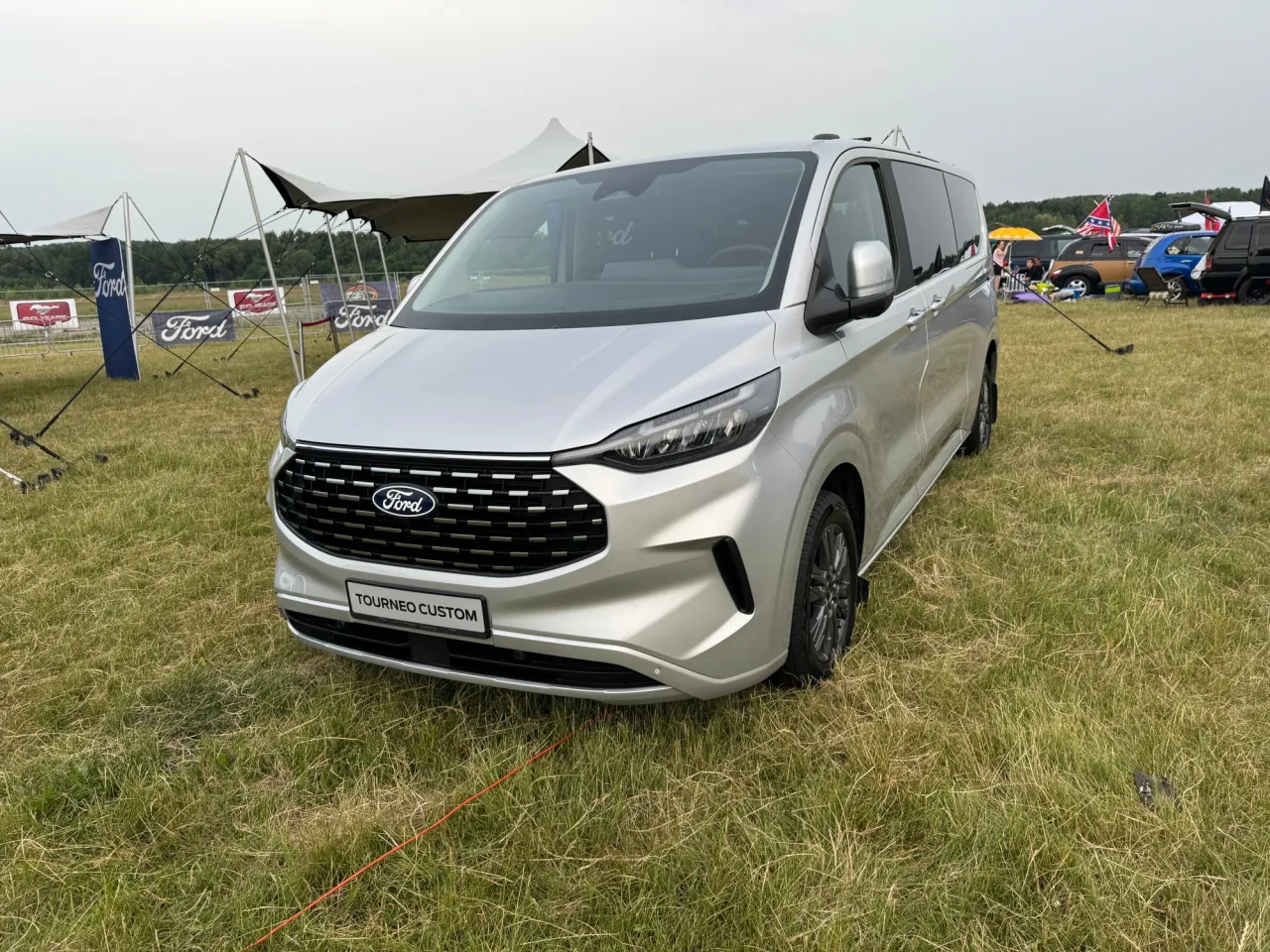 Srebrny Ford Tourneo Custom, jaki bus z napędem 4x4, stoi na trawie obok namiot&oacute;w Forda.