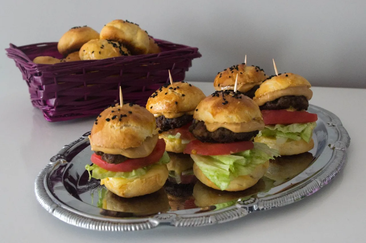 Mini bułeczki do burgerów z serem, sałatą i pomidorem na srebrnej tacy. W koszyku obok widać więcej bułeczek.