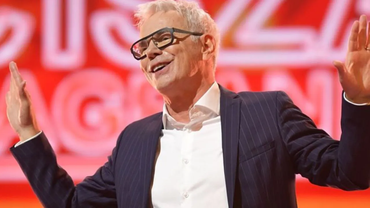 Uśmiechnięty mężczyzna w okularach, w garniturze, z rękami uniesionymi w geście. W tle czerwone logo. To pewnie komentarze do 