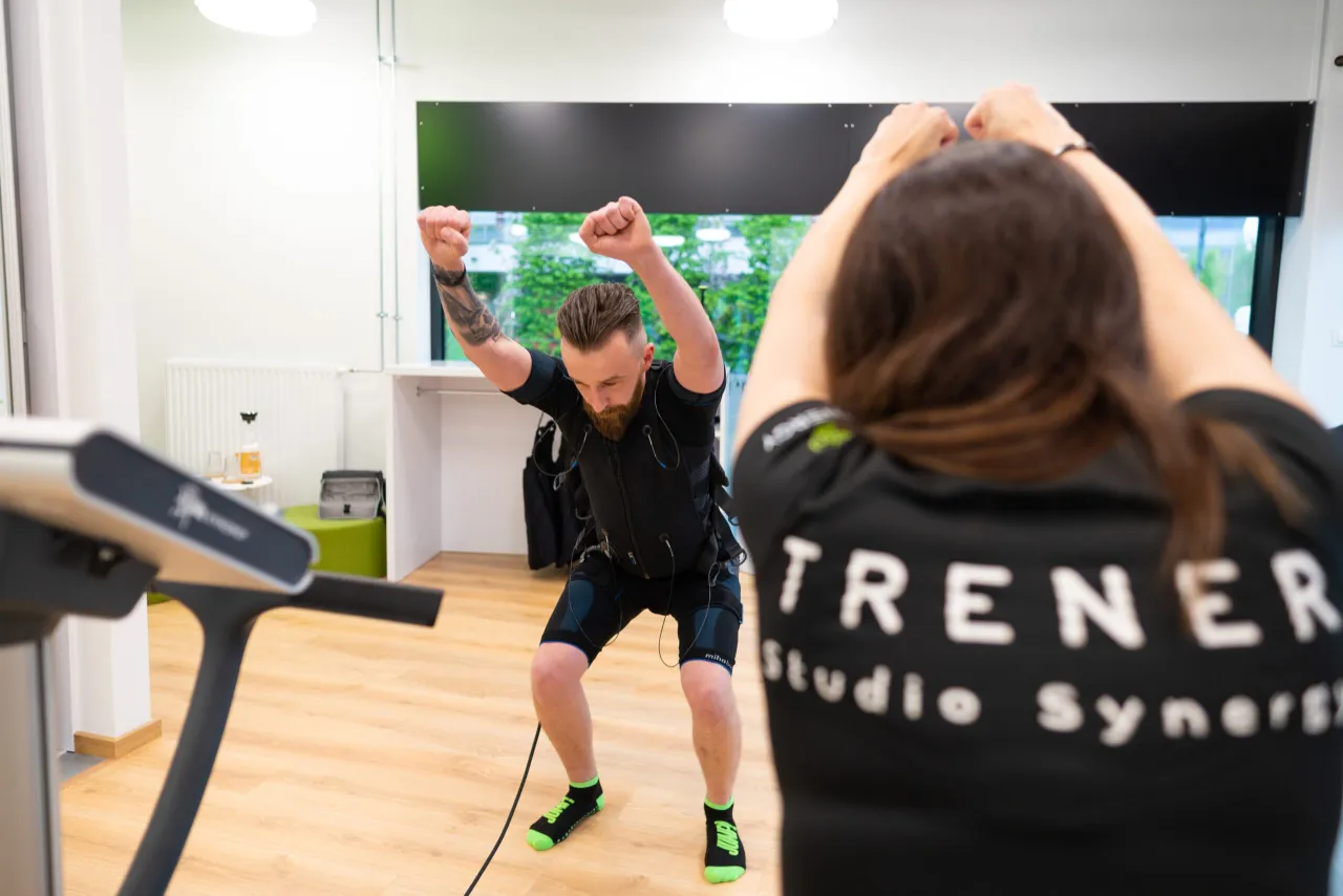 Mężczyzna w stroju EMS wykonuje ćwiczenia. Trenerka z tyłu studia patrzy na niego. Ile powinien trwać trening?