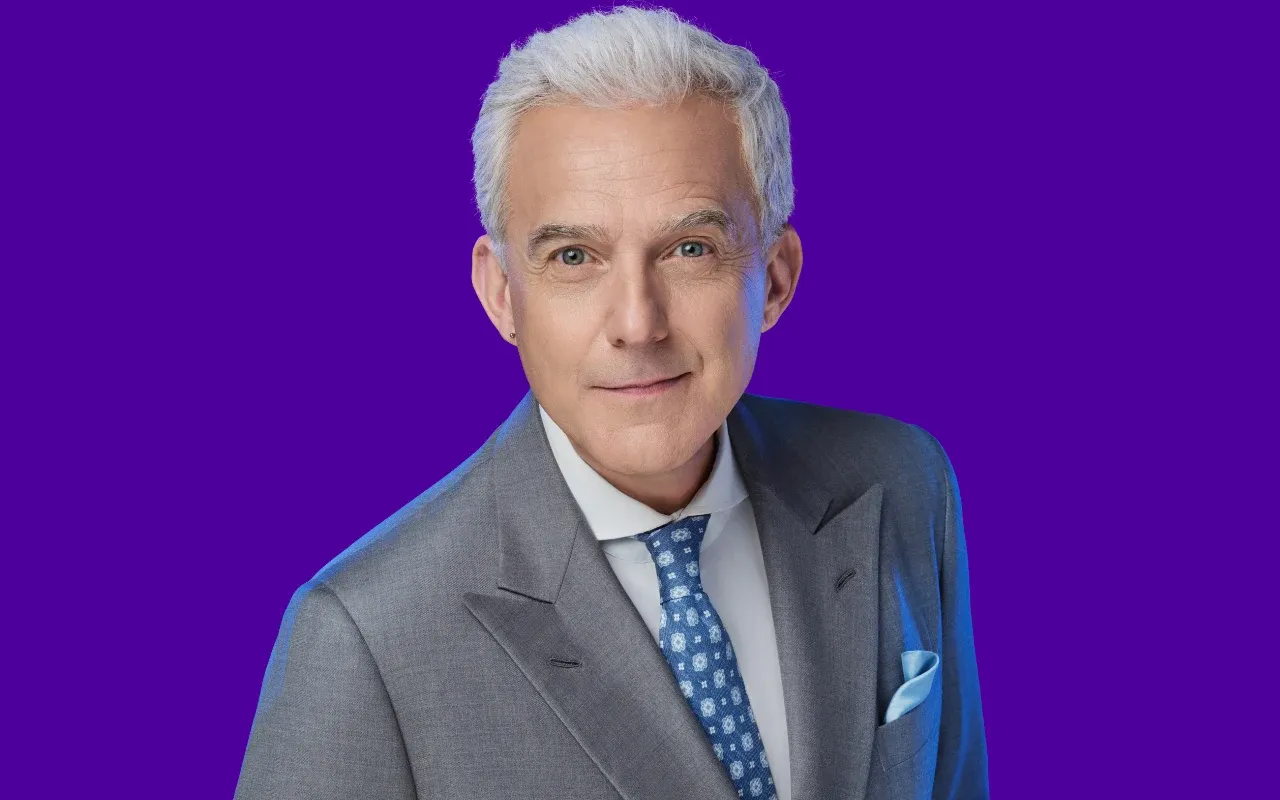 Hubert Urbański, prowadzący Milionerzy, w szarym garniturze i niebieskim krawacie.