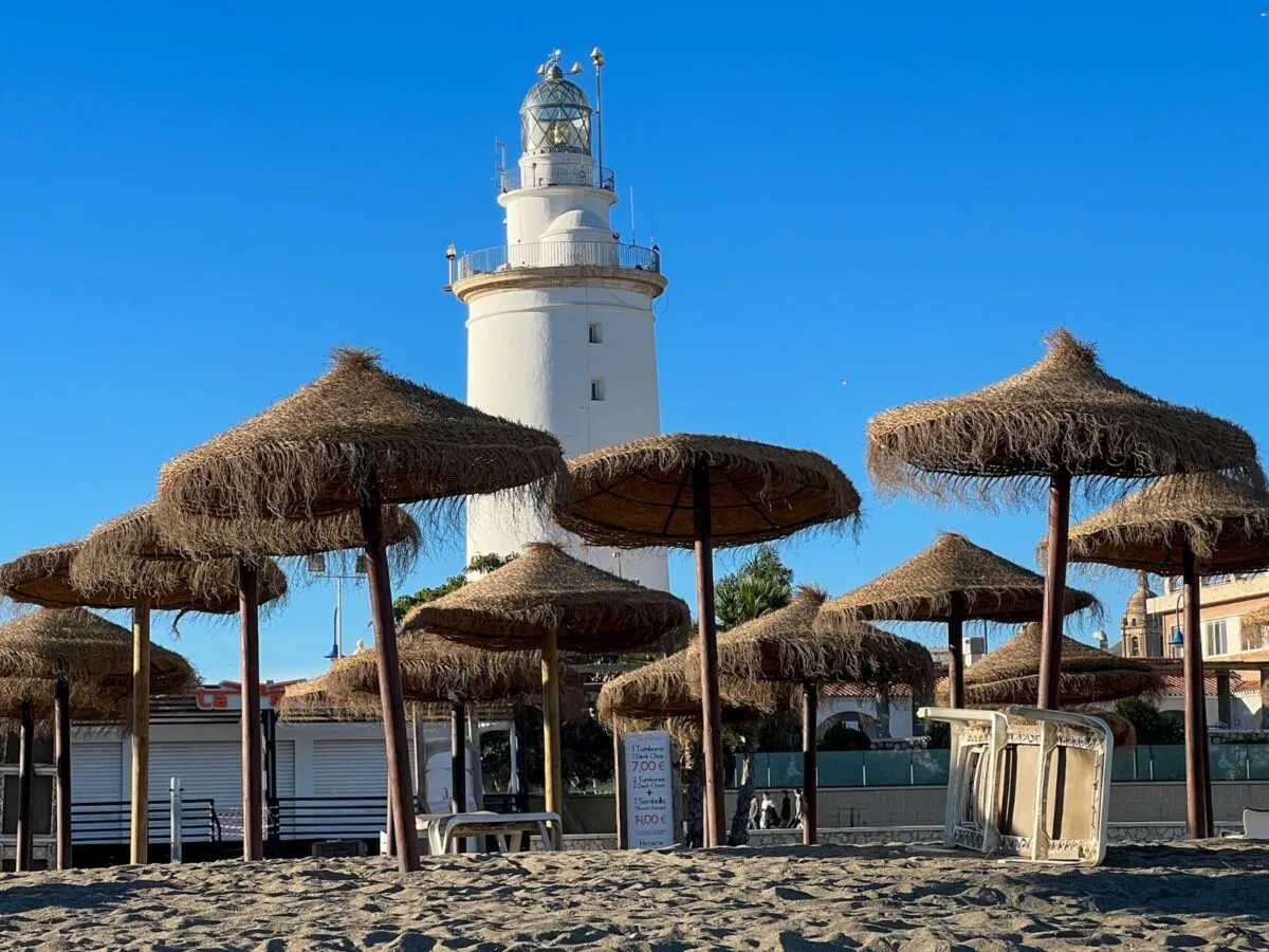 Latarnia morska góruje nad plażą z parasolami, wskazując, gdzie jest Malaga. Słońce i piasek zapraszają do odpoczynku.