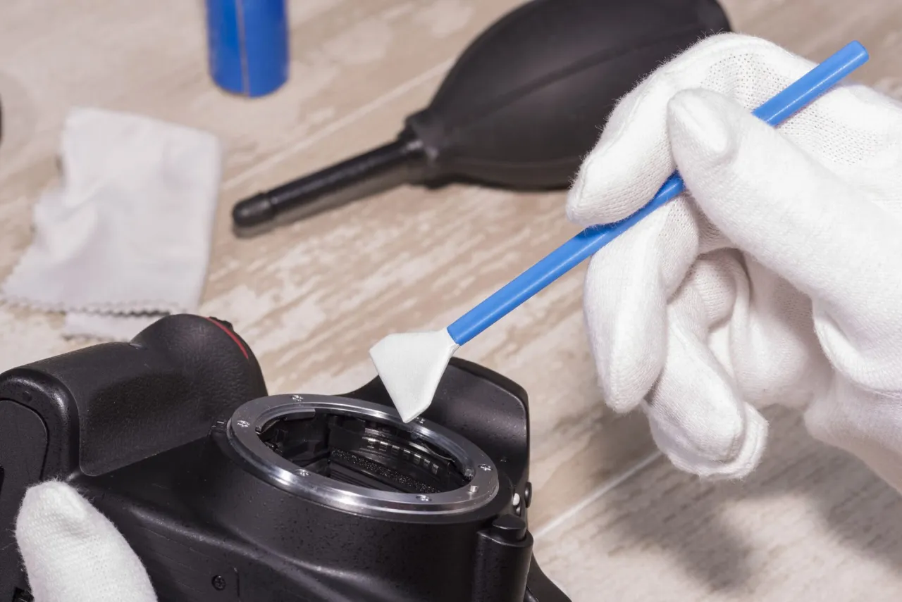 Rękawiczka w białej rękawiczce delikatnie czyści matrycę aparatu specjalnym patyczkiem. Obok leży gruszka do przedmuchiwania.