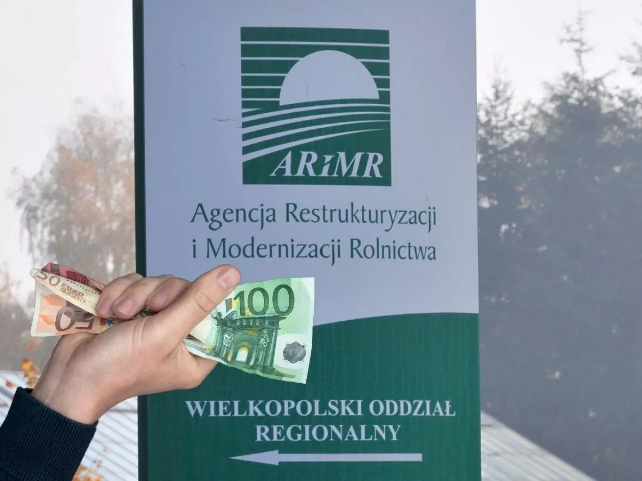Dłoń trzyma banknoty euro przed tablicą ARiMR. Chcesz wiedzieć, jak sprawdzić wysokość dopłat bezpośrednich?
