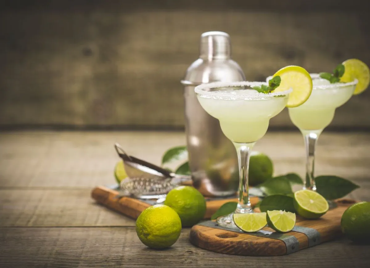Dwa orzeźwiające drinki Margarita z solą na brzegach kieliszków, udekorowane plasterkami limonki i listkami mięty. Obok shaker i świeże limonki, idealne do przygotowania Margarita przepis.