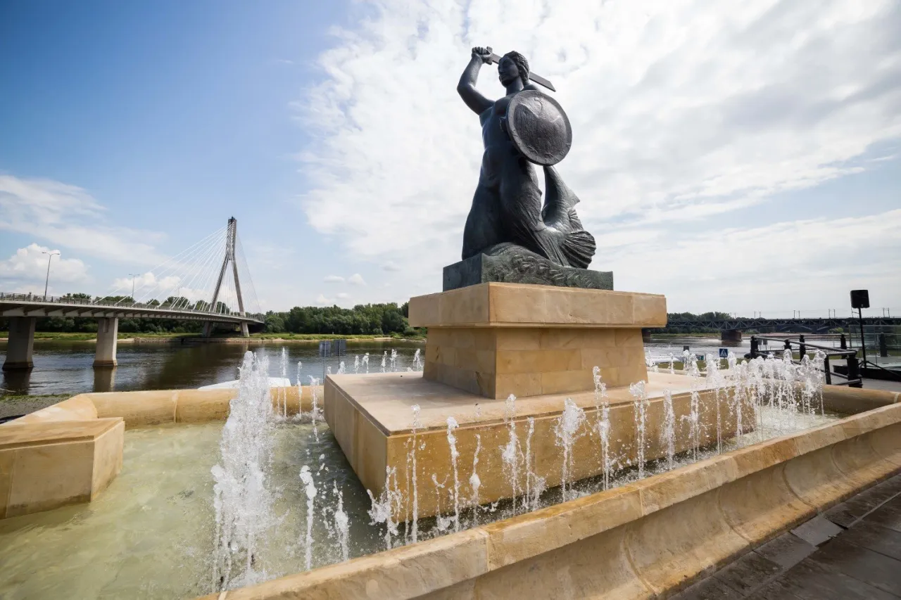 Pomnik Syrenki Warszawskiej z mieczem i tarczą, symbol legendy o syrence, na tle Wisły i mostu.