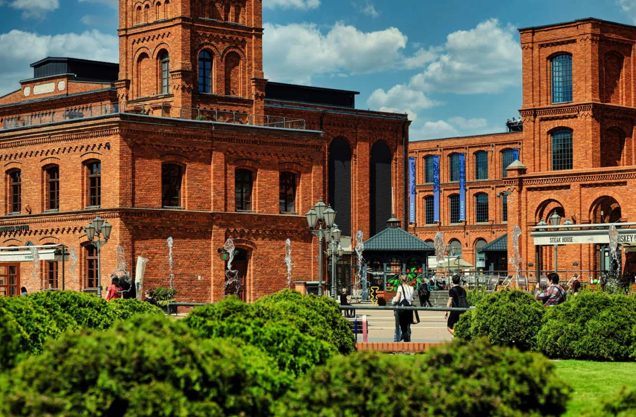Teren wok&oacute;ł muzeum sztuki w Łodzi MS2 tętni życiem. Fontanny i zielone krzewy tworzą malowniczą scenerię dla ceglanych budynk&oacute;w.