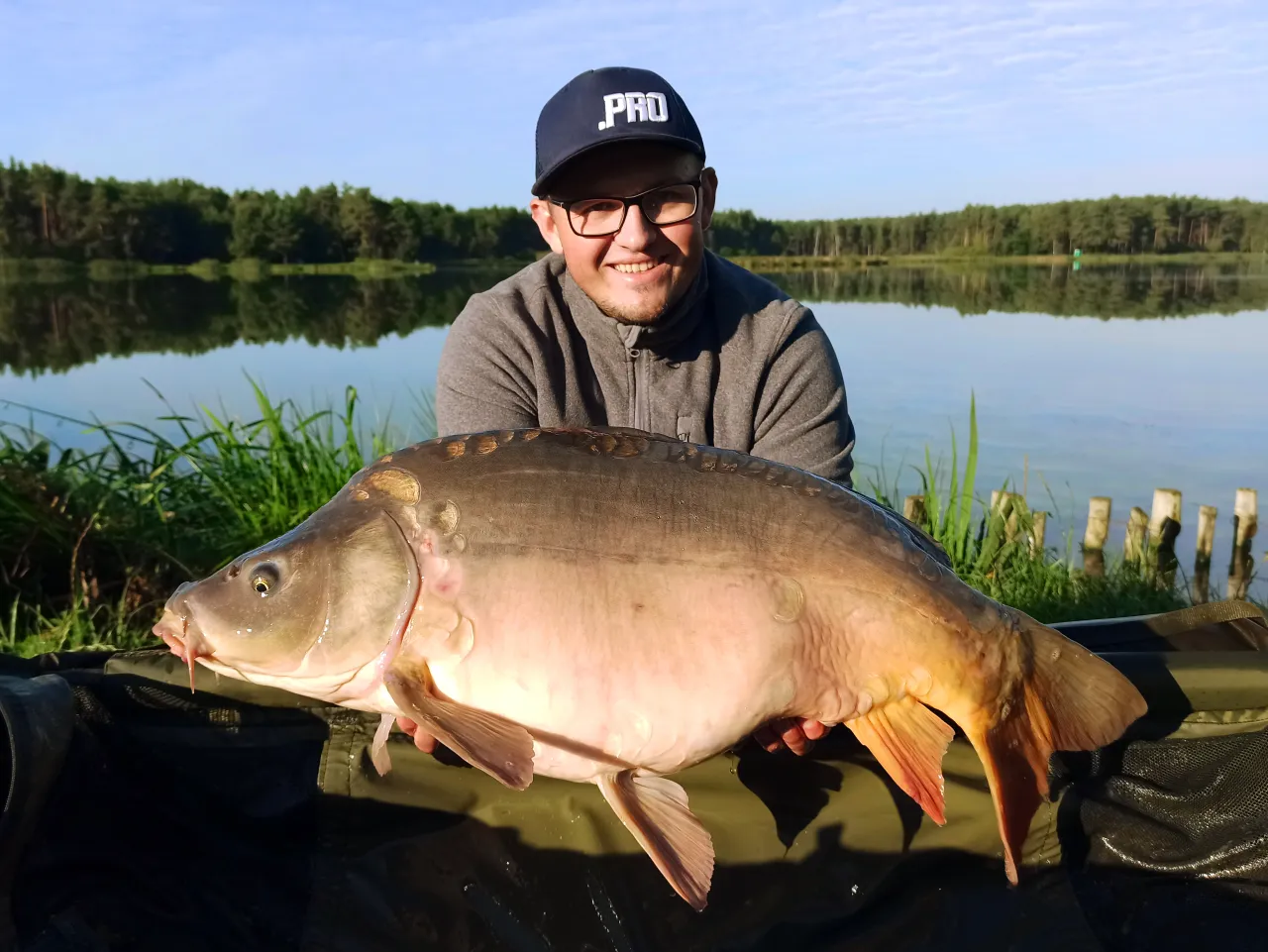 Szczęśliwy wędkarz prezentuje ogromnego karpia złowionego dzięki skutecznej zanęcie na karpia. Piękny okaz na tle jeziora.