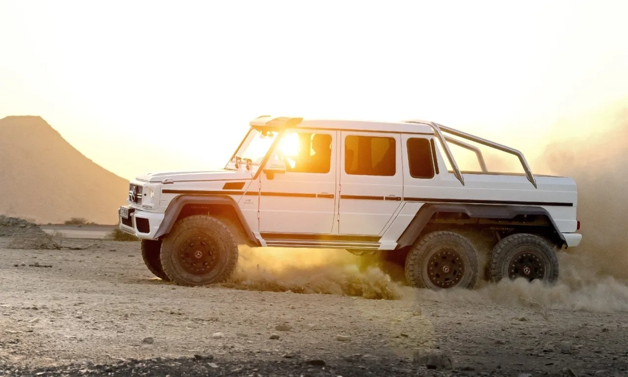 Biały Mercedes-Benz G63 AMG 6x6 szaleje po pustyni, wzbijając tumany kurzu.
