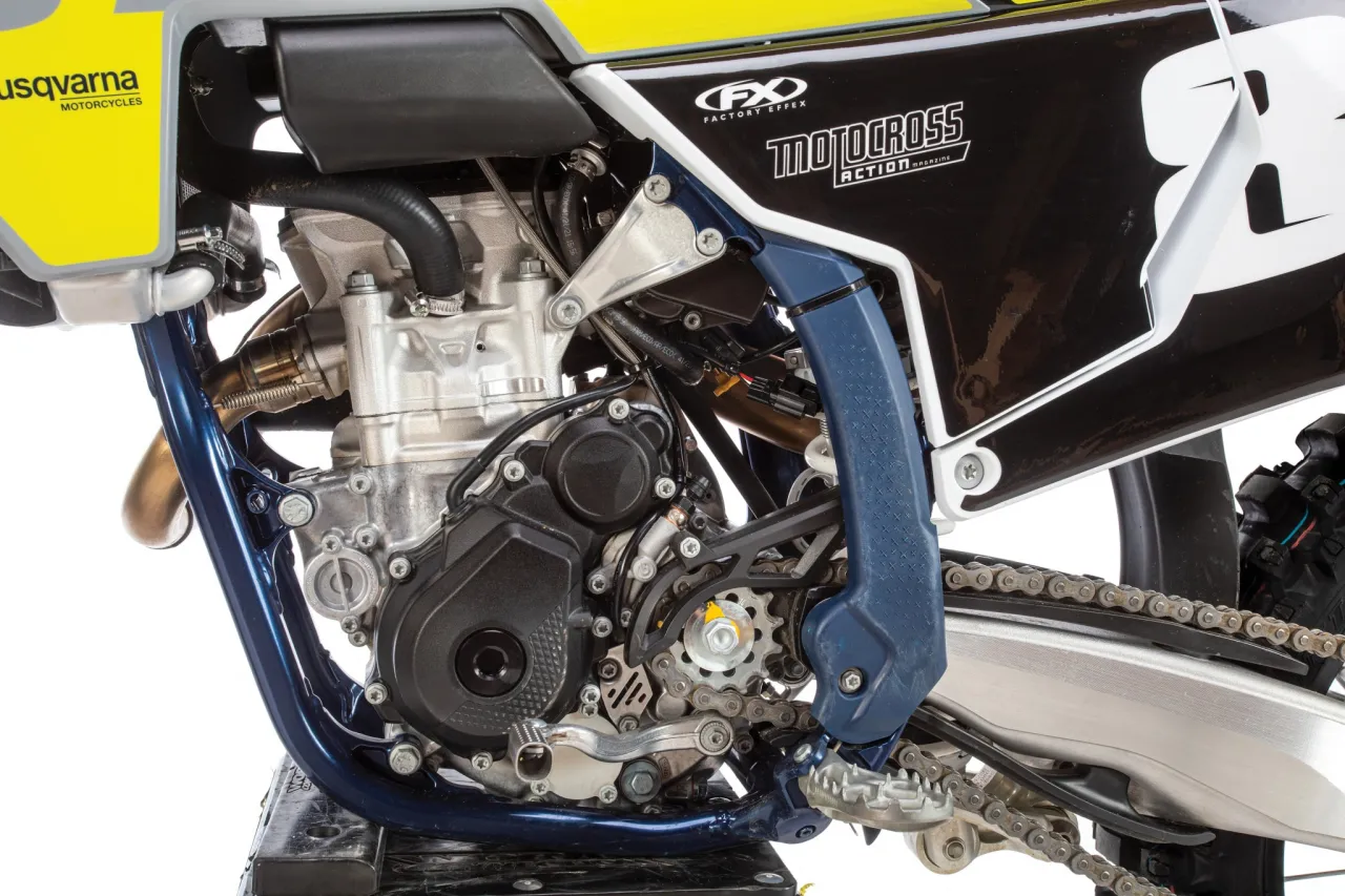 Zbliżenie na silnik i napęd motocykla Husqvarna FC 250. Widoczny jest metalowy osprzęt, łańcuch i niebieska rama.