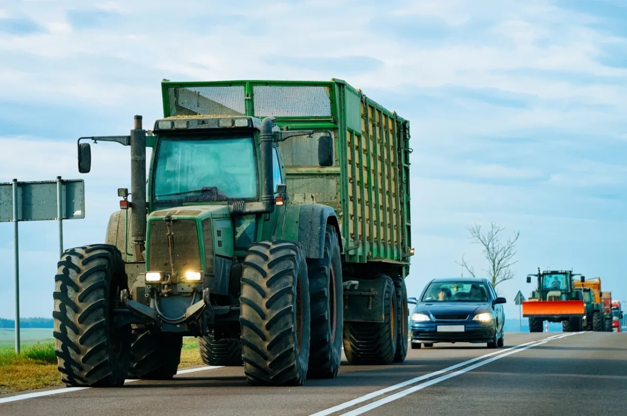 Traktor z przyczepą na drodze. Czy traktorkiem kosiarka można jeździć po drodze? Tak, widać, że maszyny rolnicze poruszają się po asfalcie.