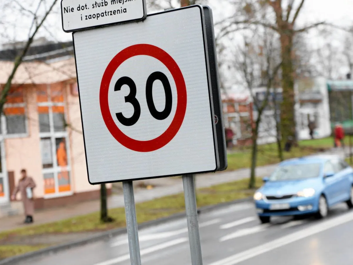 Znak ograniczenia prędkości do 30 km/h. Na drodze ekspresowej, w tle budynek i niebieski samoch&oacute;d.