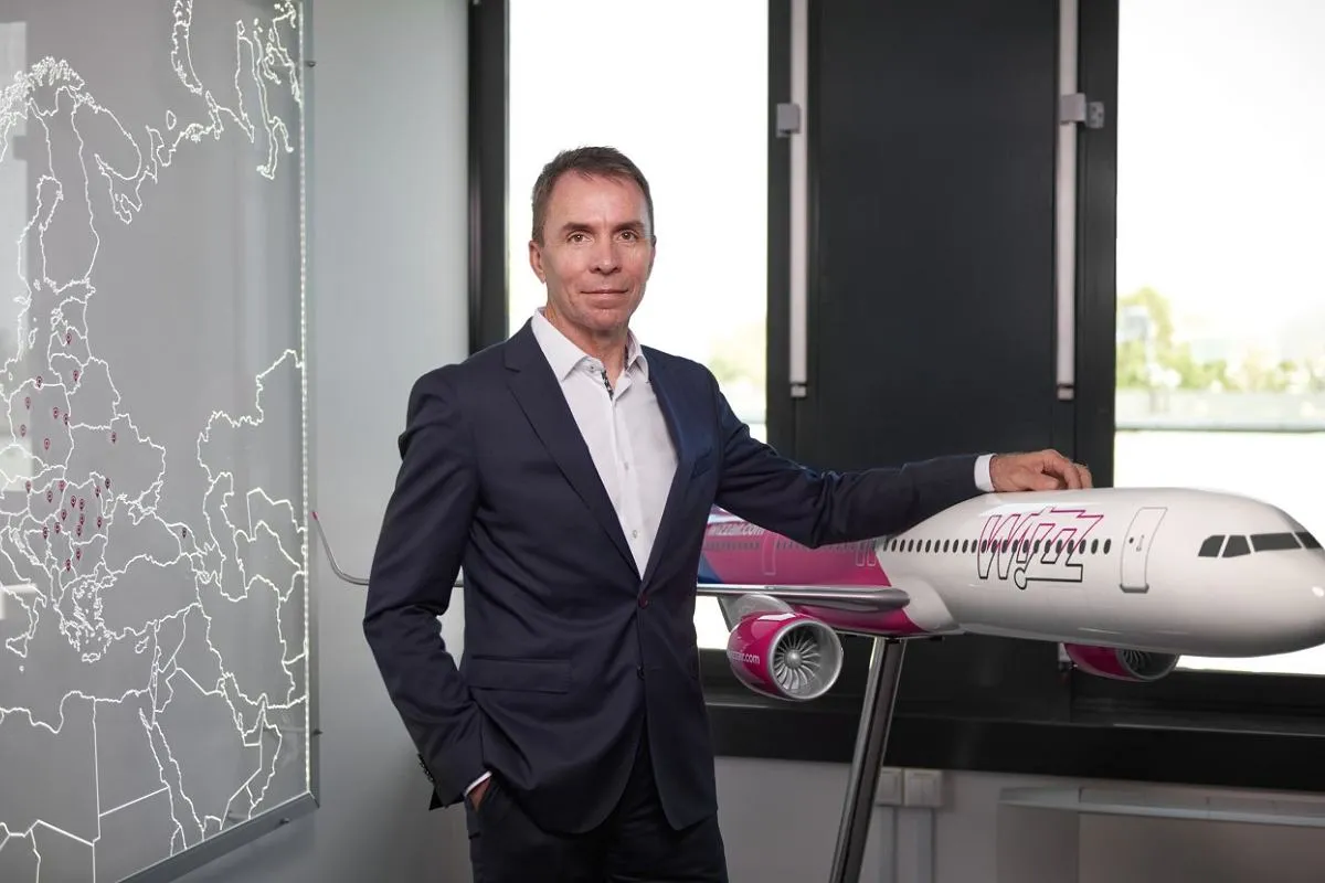 Mężczyzna w garniturze stoi obok modelu samolotu Wizz Air. Obok znajduje się mapa Europy z zaznaczonymi punktami.