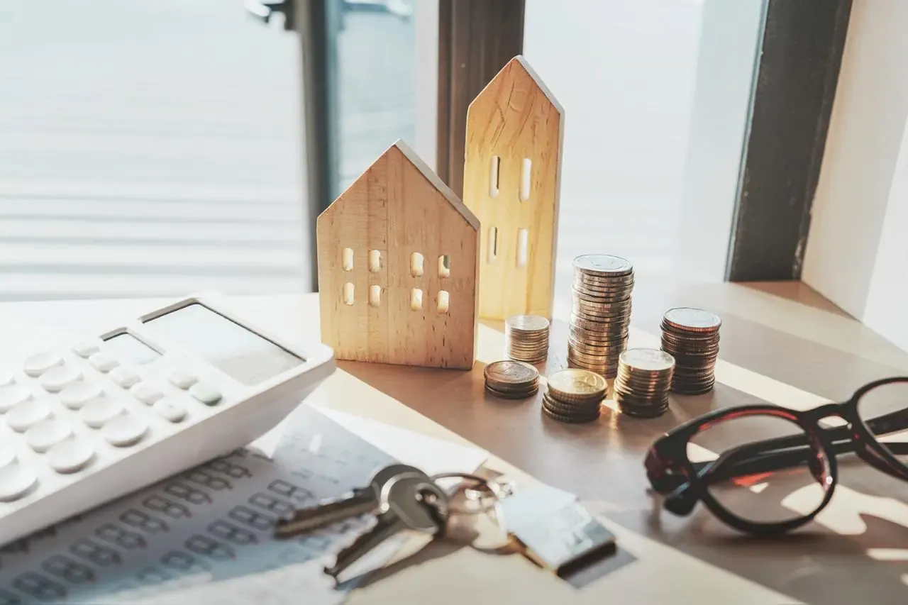 Klucze, monety i modele domów na stole. Zastanawiasz się, jak dostać kredyt na mieszkanie? Kalkulator i okulary sugerują planowanie.