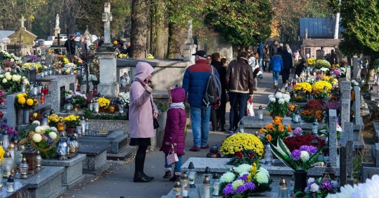 Rodzina odwiedza groby na cmentarzu, przygotowując się do Wszystkich Świętych. Widać zadbane nagrobki ozdobione kwiatami i zniczami.