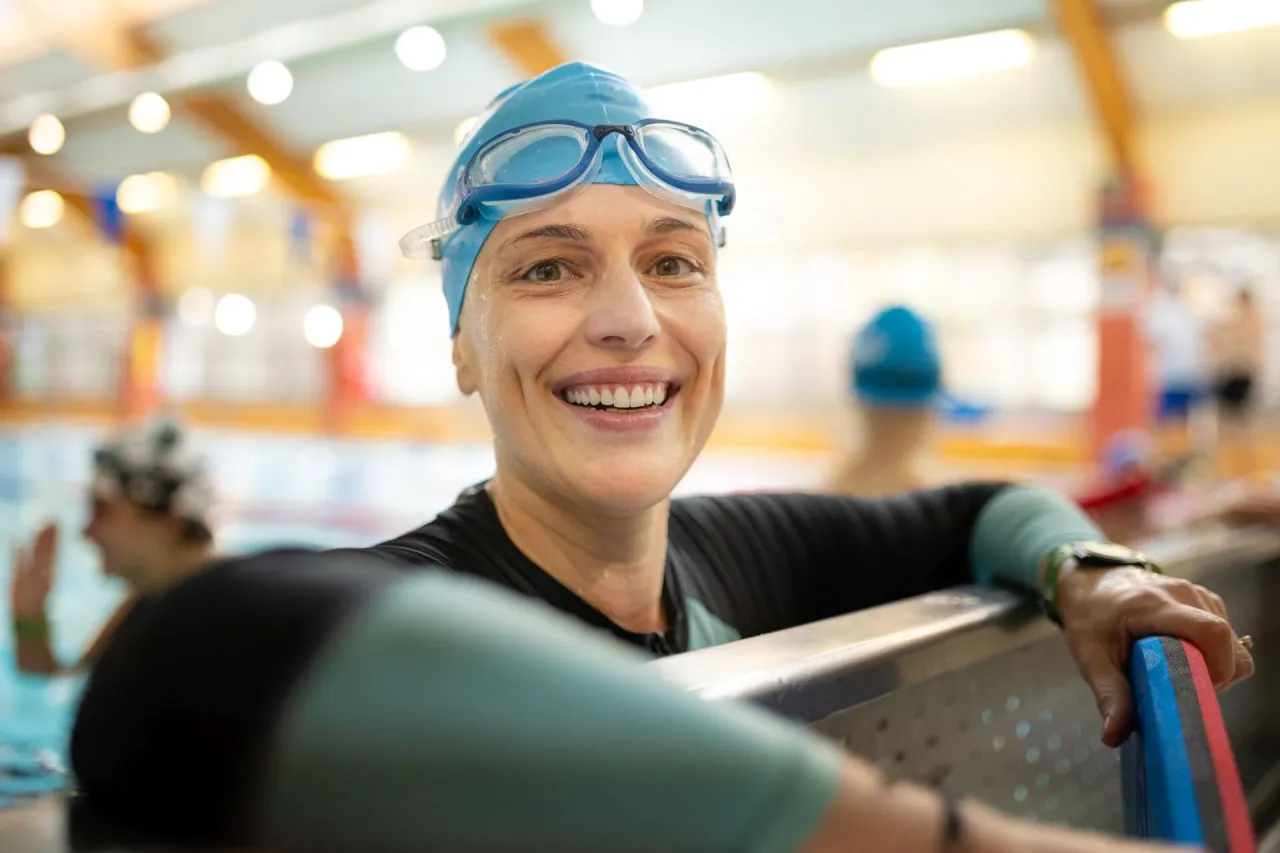 Uśmiechnięta kobieta w czepku i okularach pływackich, gotowa do nauki pływania dla dorosłych.