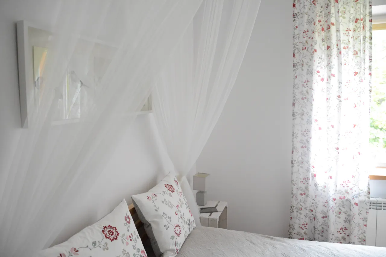 Delikatny baldachim nad łóżkiem, jak zrobić stelaż do baldachimu? Biała pościel, kwieciste poduszki i zasłony.