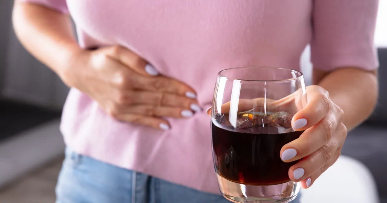 Kobieta trzyma szklankę z ciemnym napojem, odczuwając b&oacute;l brzucha po alkoholu. Co robić w takiej sytuacji?