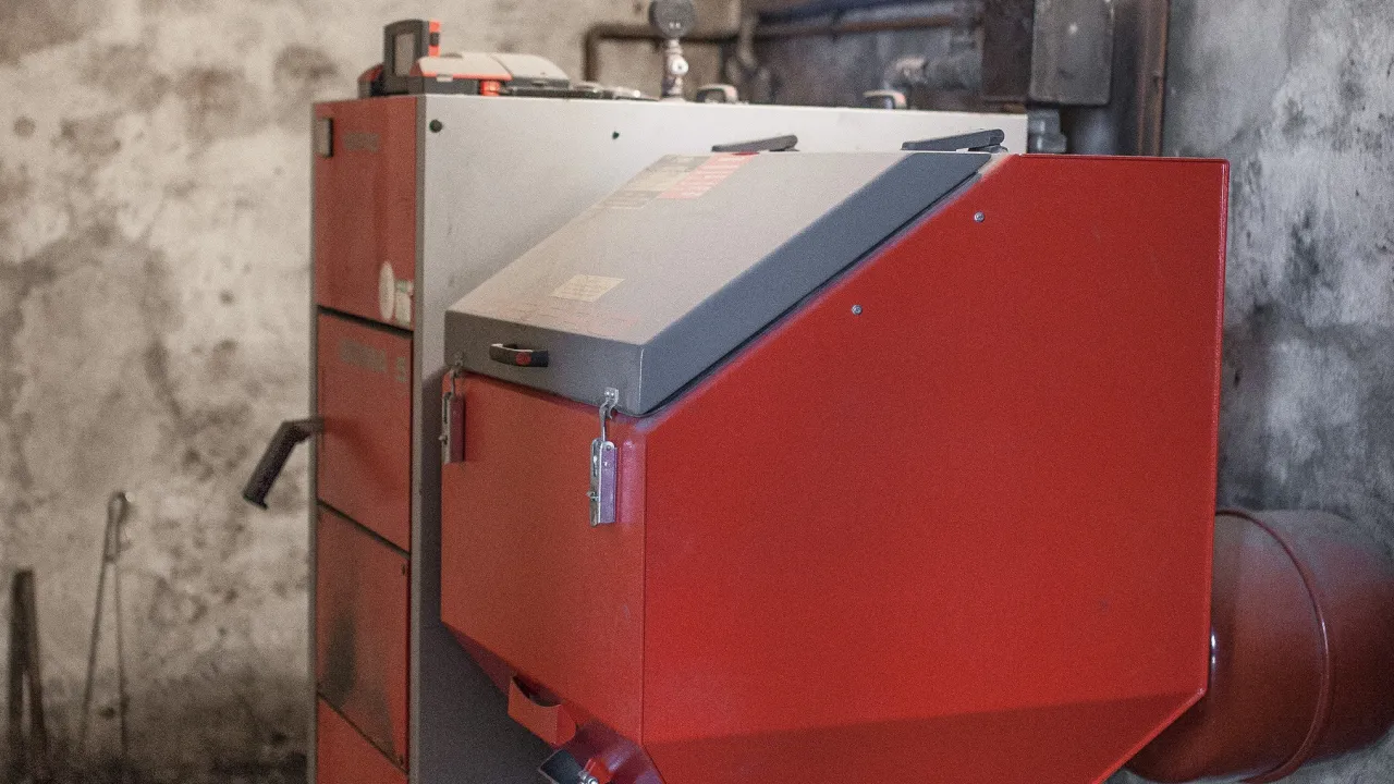Czerwony kocioł CO, prawdopodobnie klasy 5, z szarym panelem sterowania. W tle widać fragmenty instalacji i narzędzia.