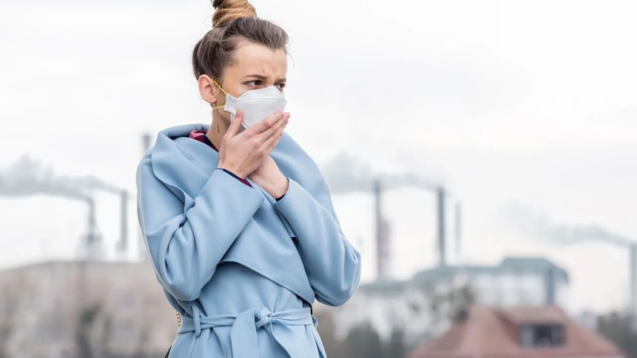 Kobieta w masce ochronnej kaszle na tle dymiących komin&oacute;w. Jak leczyć zatrucie smogiem? Unikaj zanieczyszczonego powietrza.