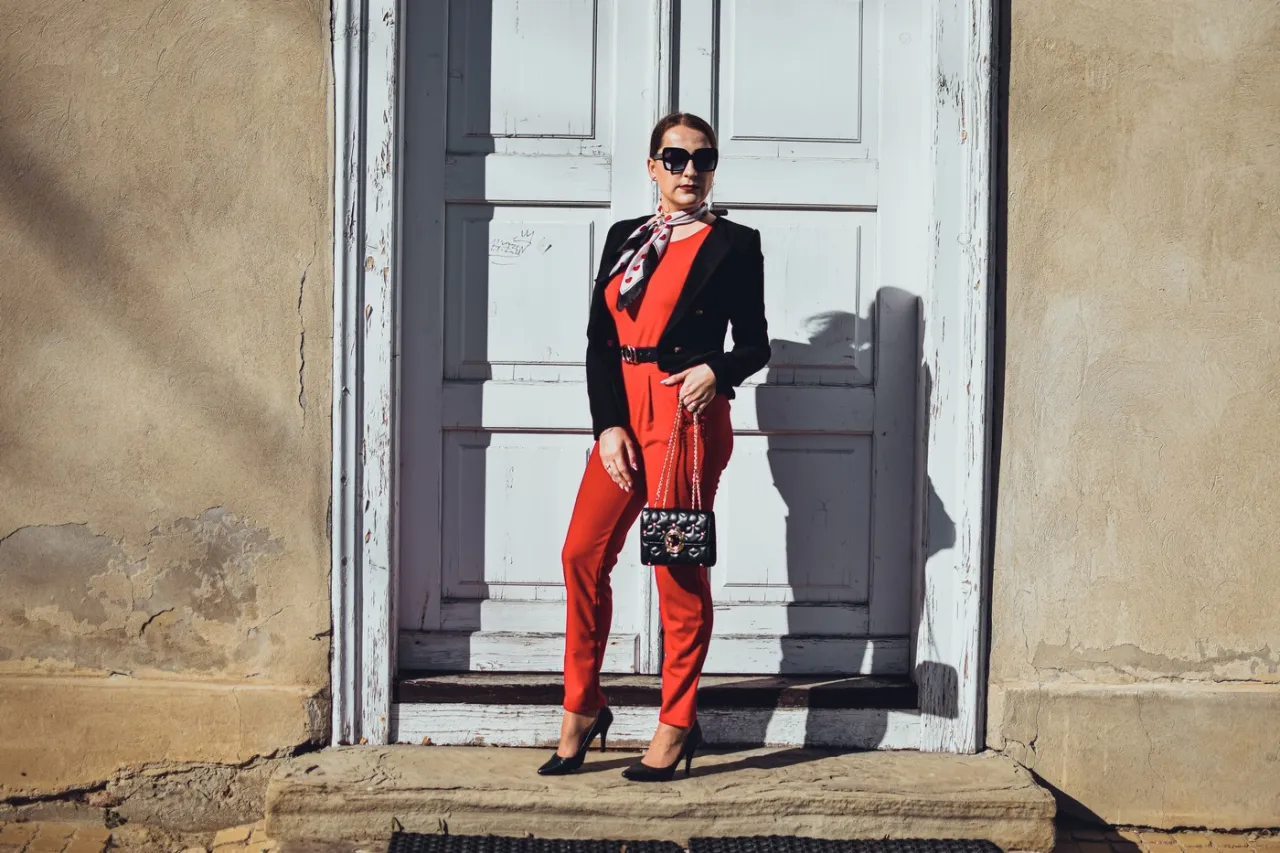 Elegancka kobieta w czerwonym kombinezonie i czarnym żakiecie, zastanawia się, jakie buty do czerwonego kombinezonu będą najlepsze. Cz&oacute;łenka idealnie pasują.