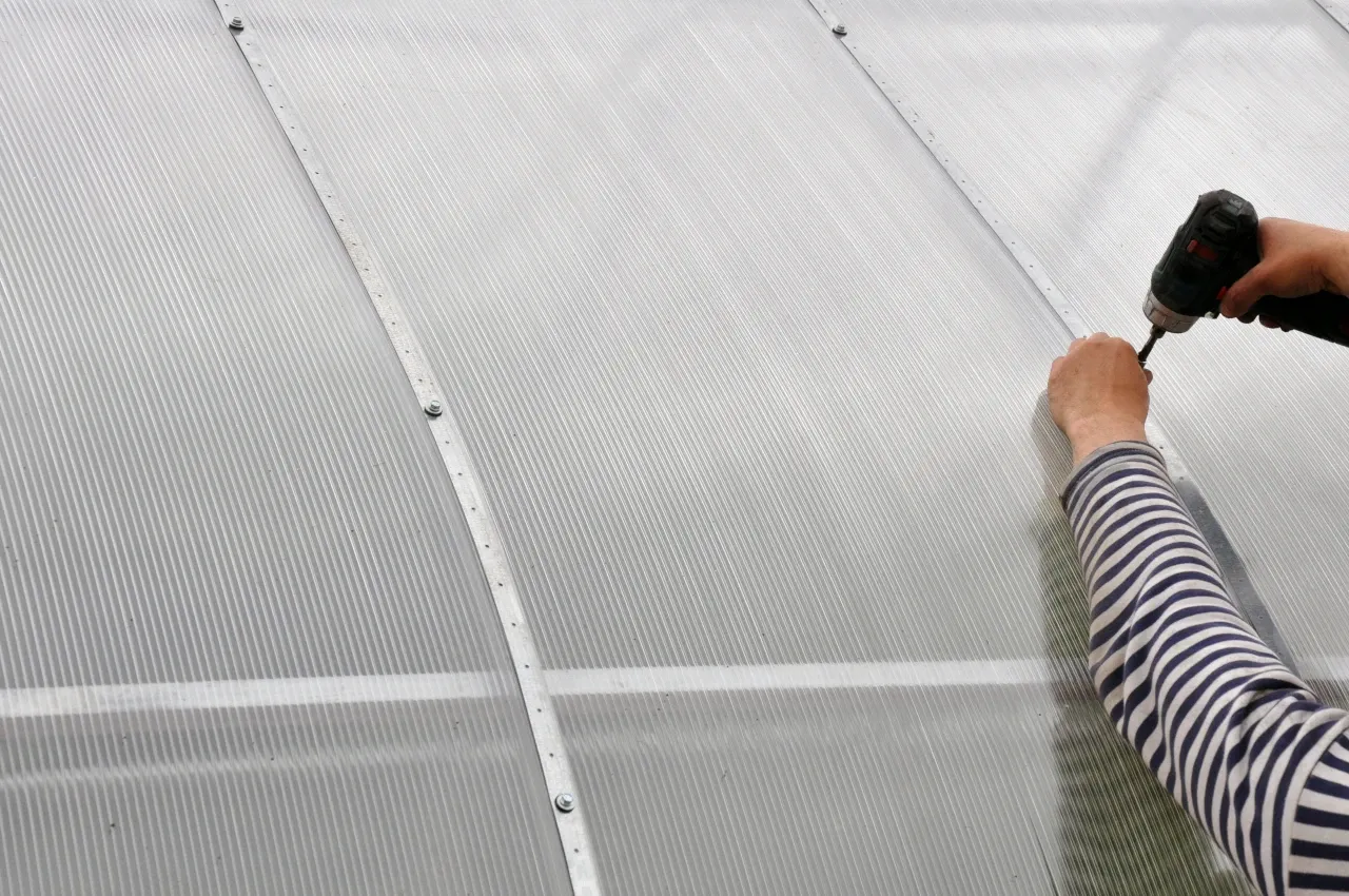 Montaż szklarni z poliwęglanu: ręka z wkrętarką przykręca panel.