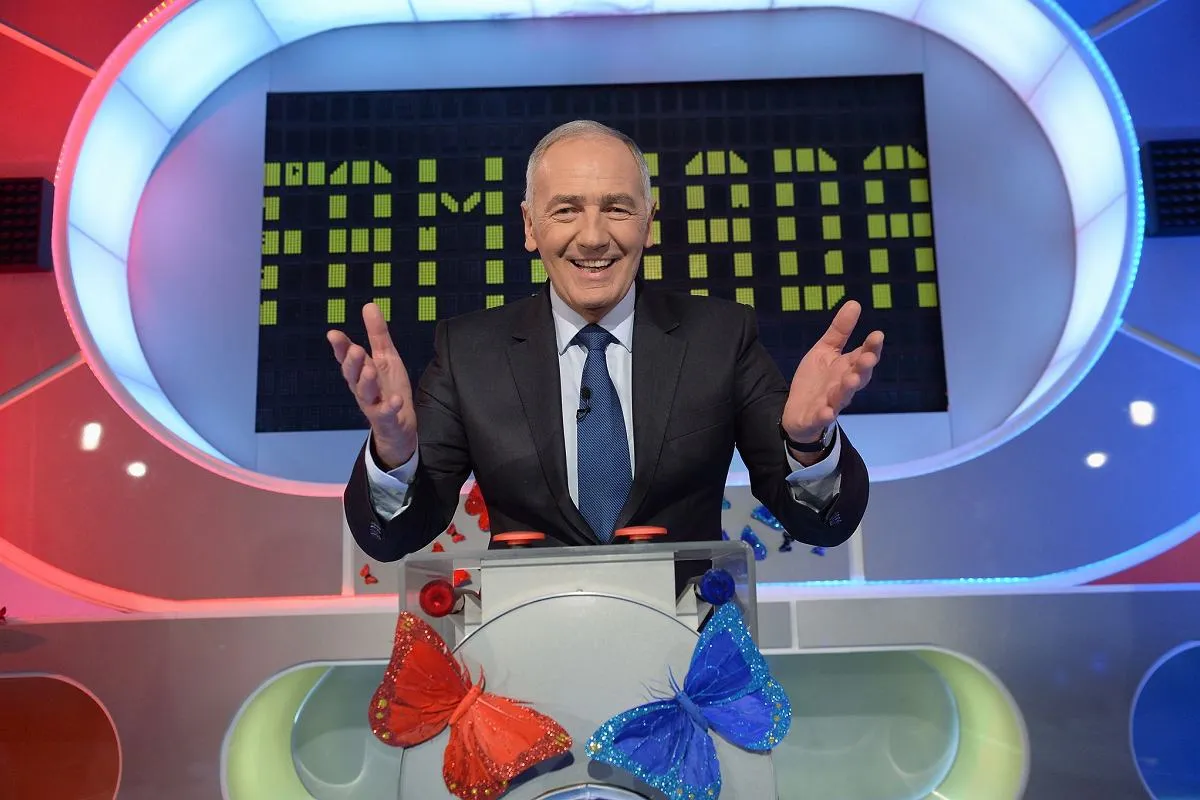 Uśmiechnięty mężczyzna w garniturze, prowadzący teleturniej, z motylami na panelu. Taki może być pilot Familiady.