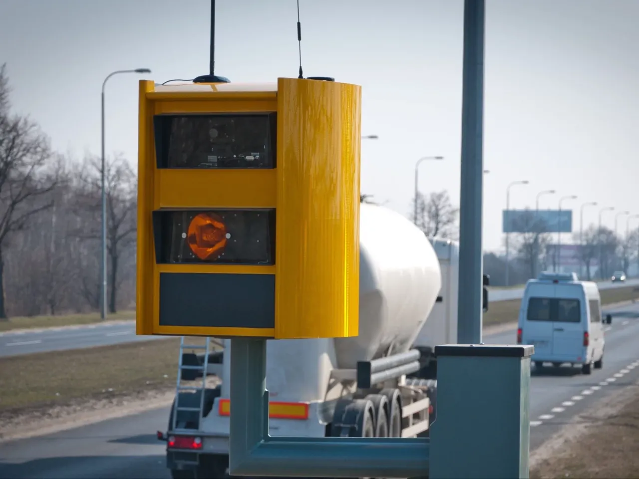 Ż&oacute;łty fotoradar przy drodze, obok ciężar&oacute;wka i biały bus. Zastanawiasz się, po jakim czasie przychodzi mandat z fotoradaru?