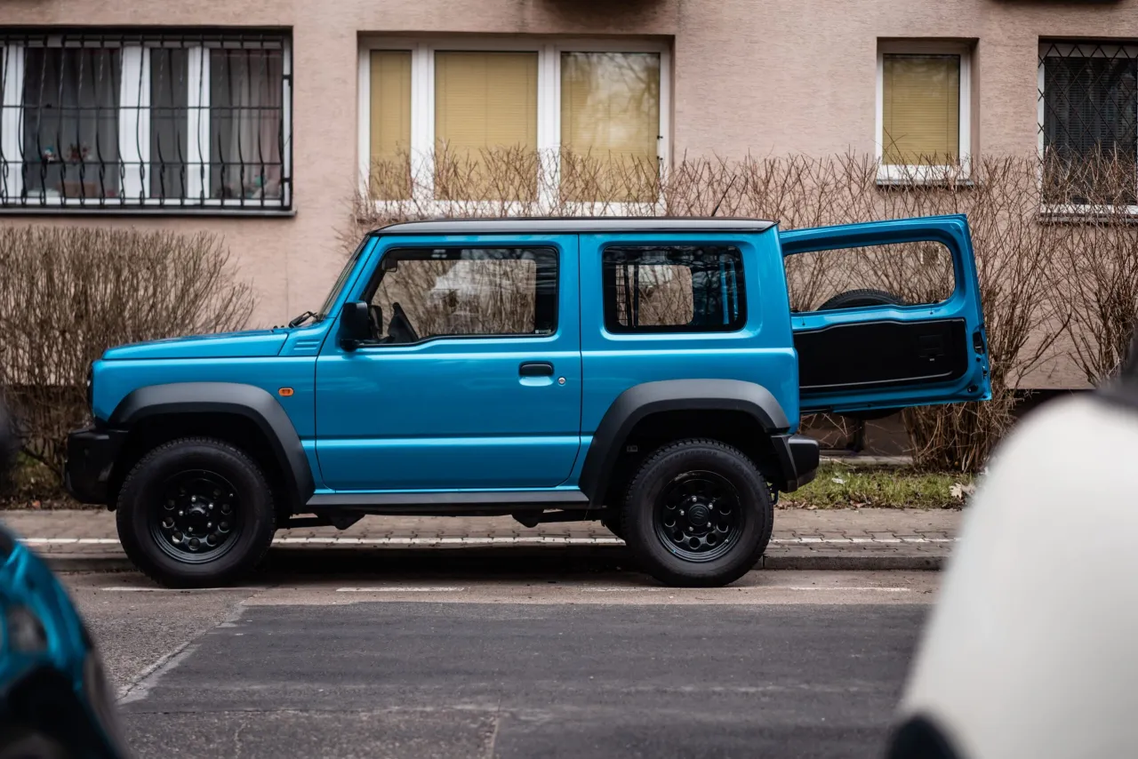 Niebieski Suzuki Jimny z otwartymi tylnymi drzwiami, gotowy do podróży. Jego pojemność baku pozwala na długie wyprawy.