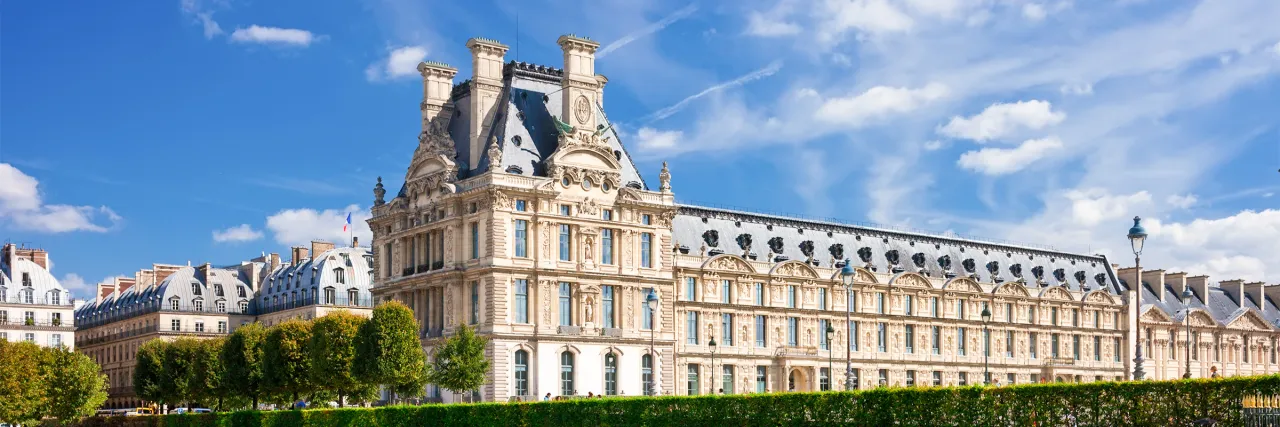 Elegancka fasada budynku w 1 dzielnicy Paryża, z ozdobnymi detalami i mansardowym dachem, na tle błękitnego nieba z chmurami.