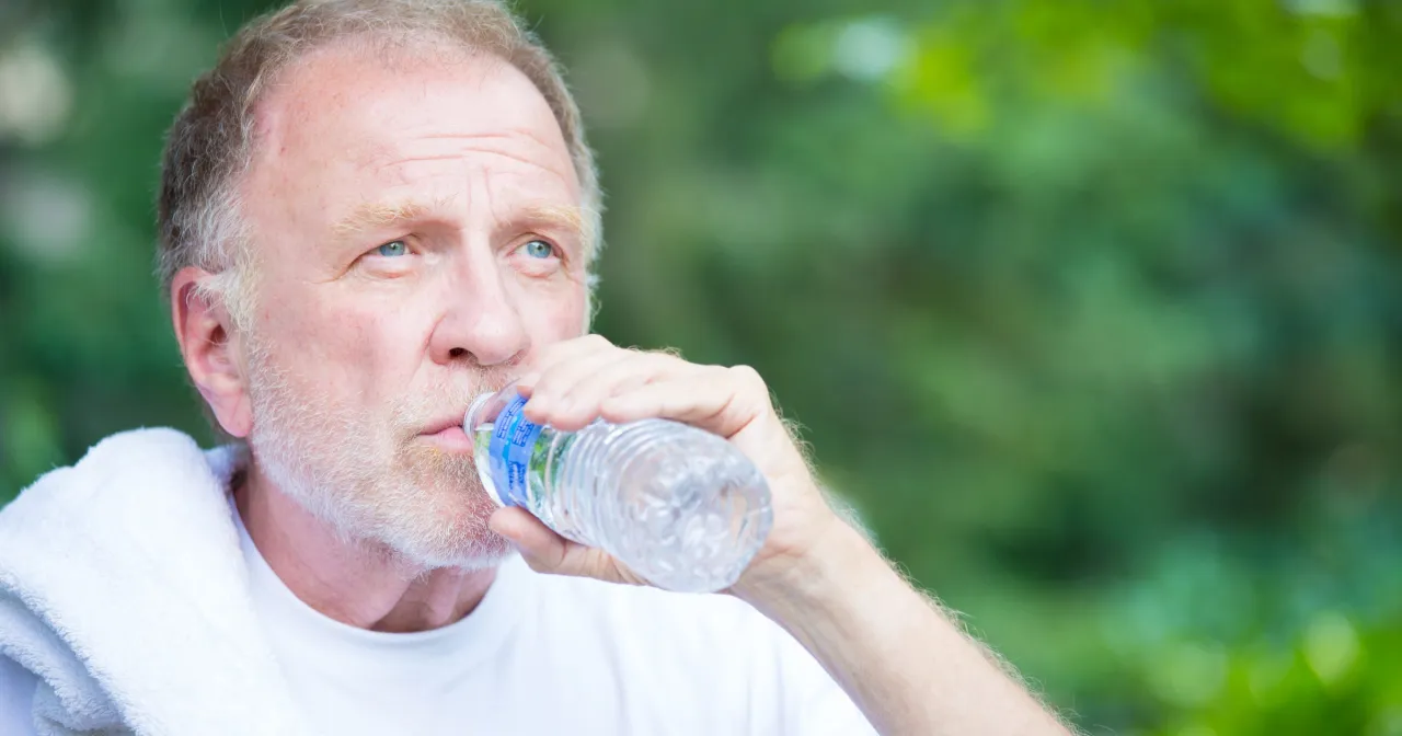 Starszy mężczyzna z ręcznikiem na ramieniu pije wodę z butelki. Może to być oznaka objawów odwodnienia u starszej osoby.