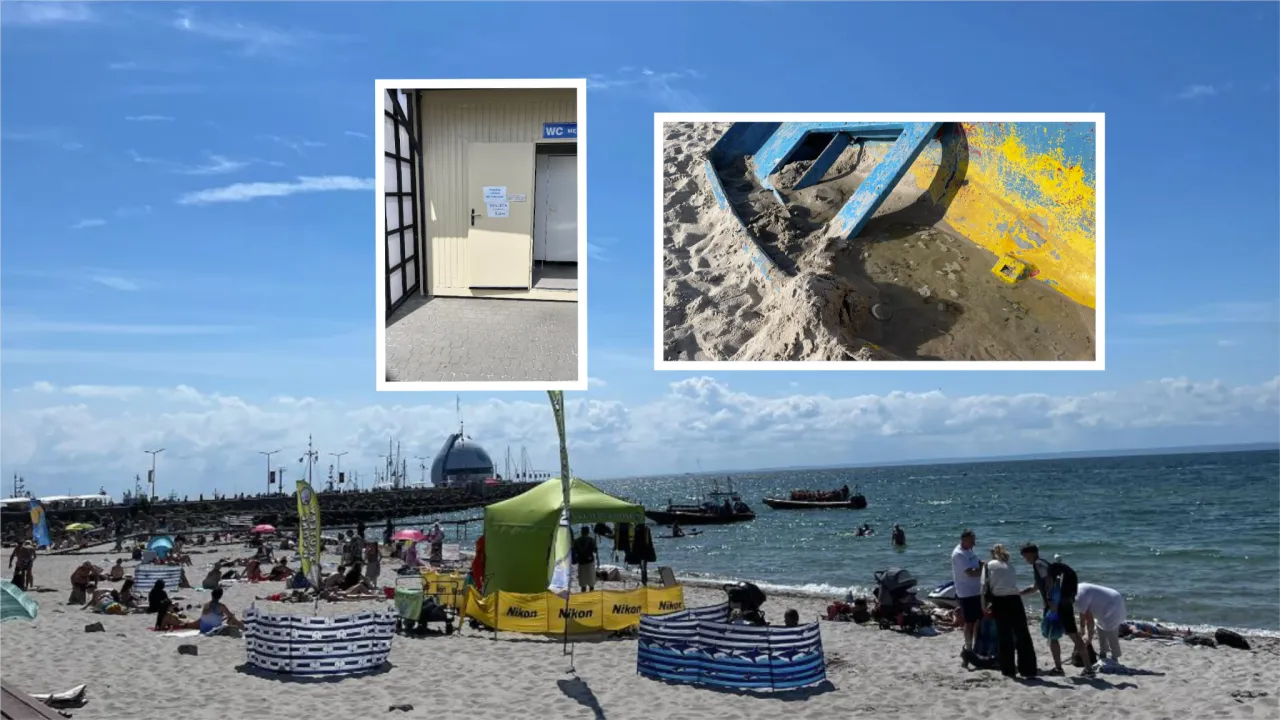 Słoneczny dzień na plaży w jednym z nadmorskich miast. W oddali widać budynek toalet i fragment wraku łodzi na piasku.