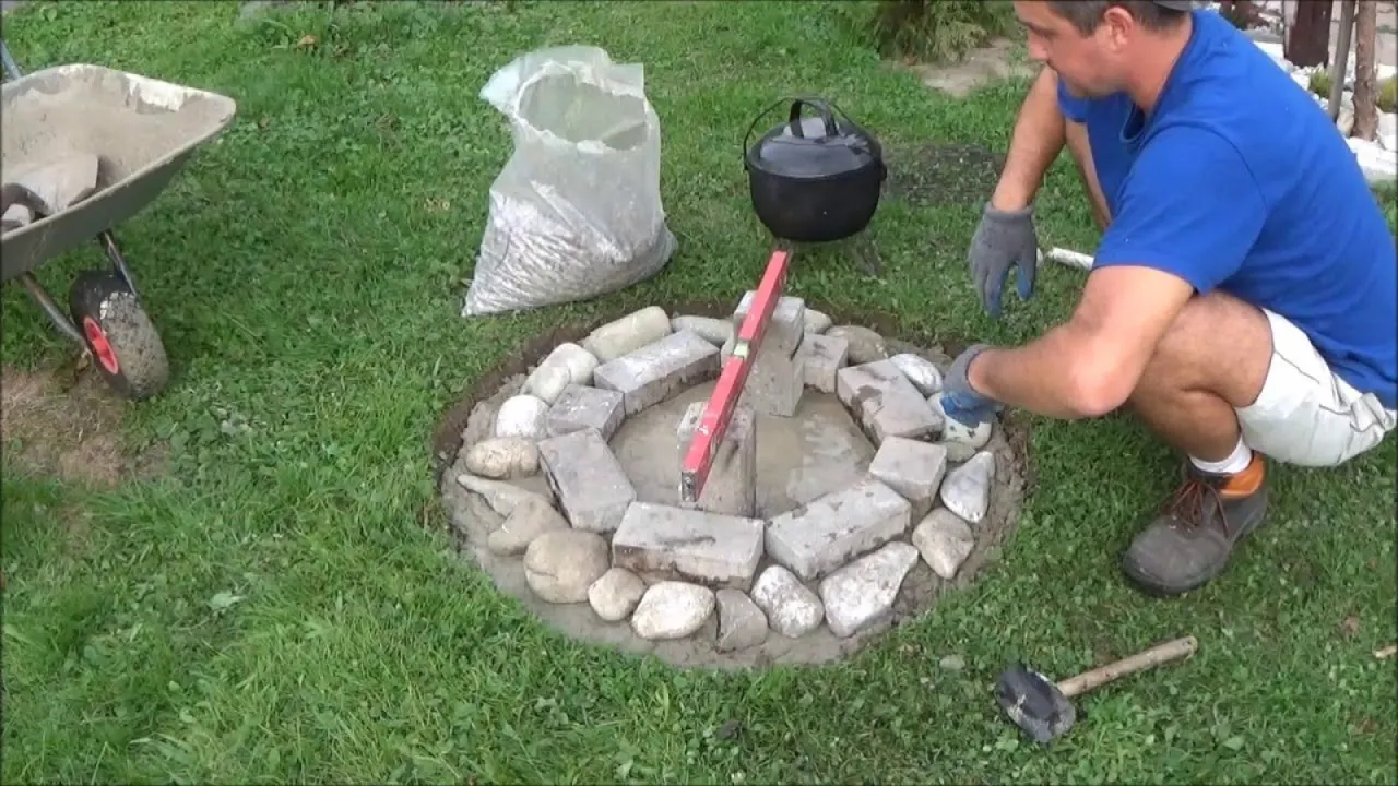 Budowa paleniska ogrodowego z kamienia: mężczyzna układa kamienie, używa poziomicy. W tle wózek i worek z kruszywem.