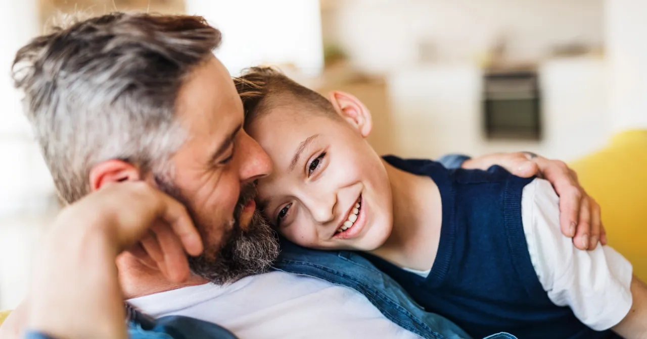 Uśmiechnięty chłopiec opiera głowę na ramieniu ojca. W tle widać fragmenty domu. To obraz więzi i mądrości, jak w cytaty ojca do syna.
