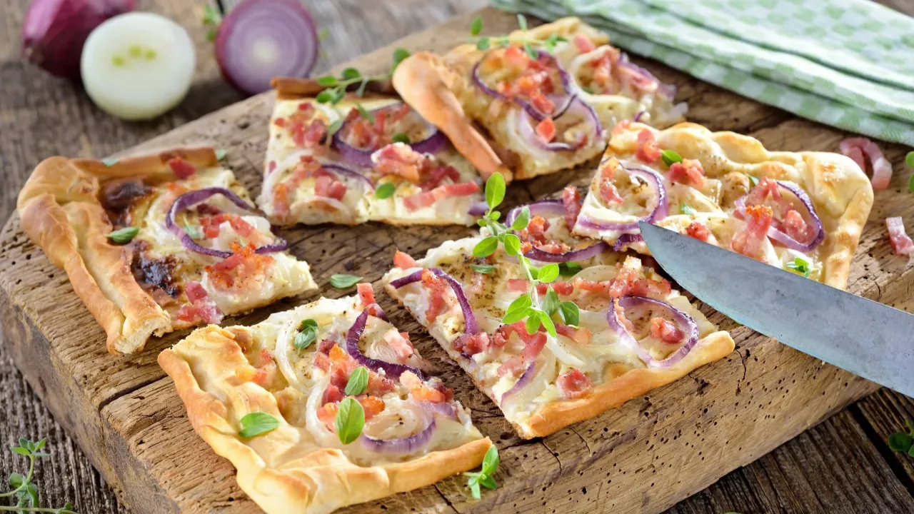 Gotowe ciasto na pizzę z cebulą i boczkiem, pokrojone na prostokątne kawałki, leży na drewnianej desce.