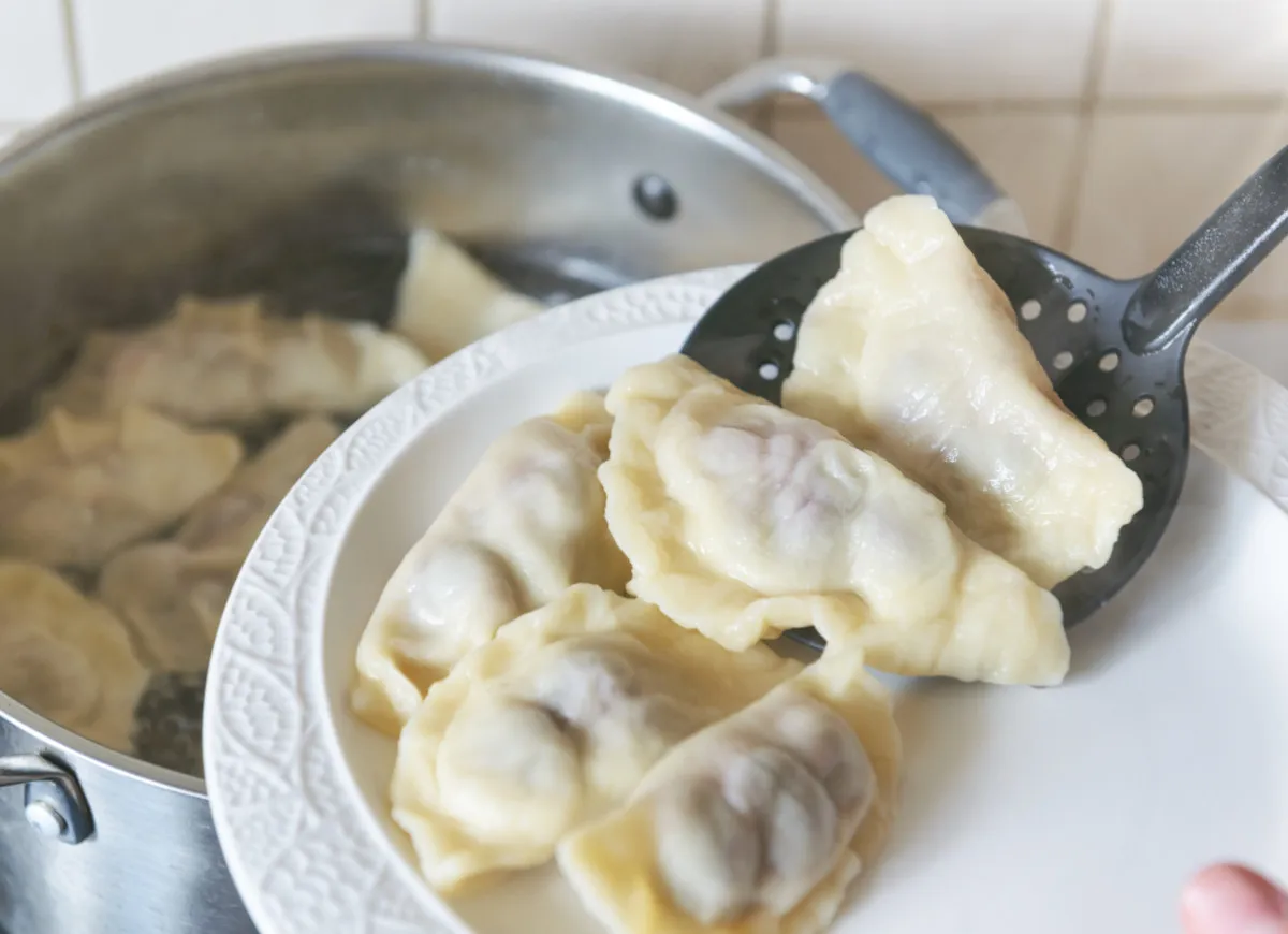 Gotowanie pierog&oacute;w: świeżo ugotowane pierogi wyjmowane łyżką cedzakową z garnka na biały talerz.