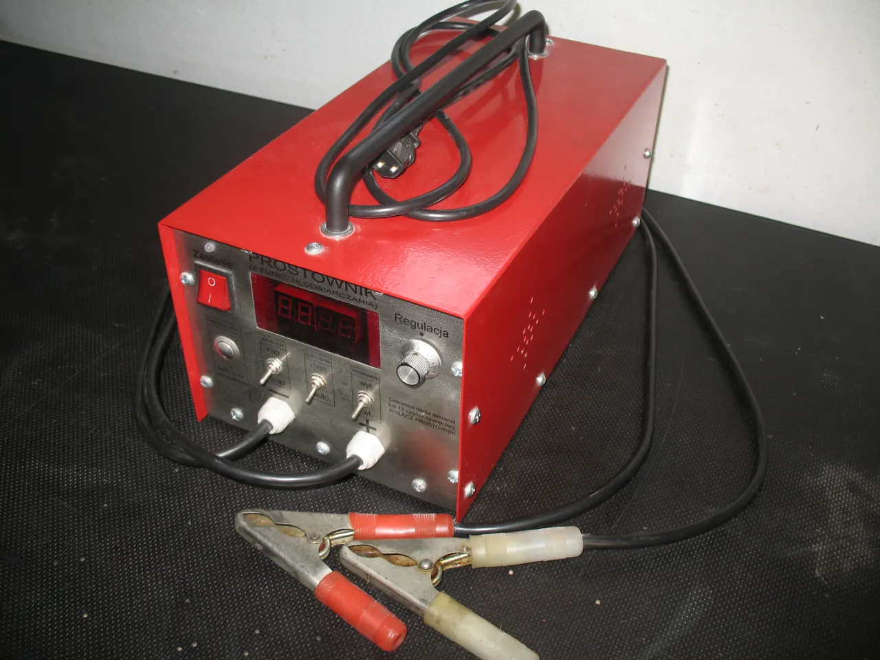 Czerwony prostownik z funkcją odsiarczania. Na panelu cyfrowy wskaźnik i przełączniki. Kable z krokodylkami.